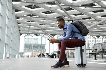 young black man sitting on suitcase at station with mobile phone