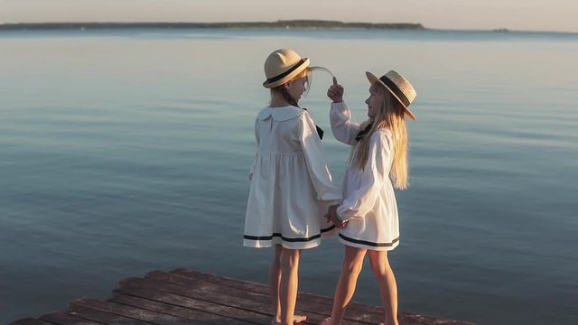 Happy girls in beautiful dresses are playing on the sea pier. In the background sea sunset. Wooden pier