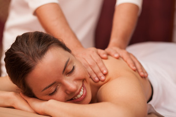 Fototapeta na wymiar Attractive joyful young woman smiling cheerfully why professional masseur massaging her shoulder. Gorgeous woman relaxing at day spa. Therapist giving soothing massage to female client. Beauty concept