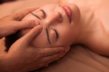 Close up of a happy beautiful woman smiling with her eyes closed, receiving soothing head massage. Young attractive woman enjoying face massage at spa center. Skincare, health, pampering concept
