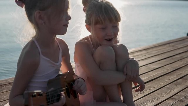 Beautiful girls play the guitar and sing funny songs. Children play on the sea coast. Evening time, calm sea.
