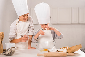 smiling brother and sister in aprons and chef hats during food preparation at table in kitchen