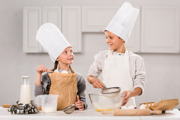 little children in aprons and chef hats preparing at table in kitchen