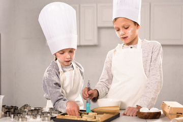 concentrated kids in aprons brushing cookies on baking tray in kitchen