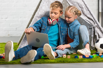 adorable little brother and sister using laptop near tent at home