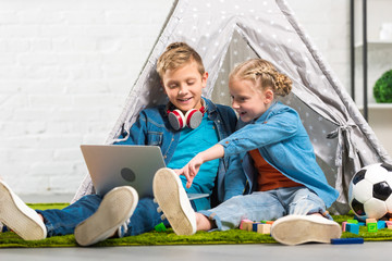 child pointing at laptop screen to smiling brother with headphones over neck near tent at home