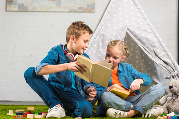 boy with headphones over neck showing book to little sister near wigwam at home © LIGHTFIELD STUDIOS