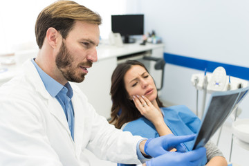 Obraz premium Woman holding her hurtful tooth having painful face expression sitting at the dentist office while beautiful young successful dentist is holding and looking at the teeth x-ray.