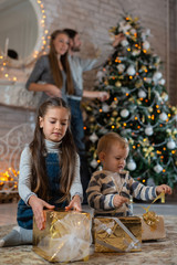 Sister hugs little brother sitting on the floor with gifts on background of their parents and Christmas tree