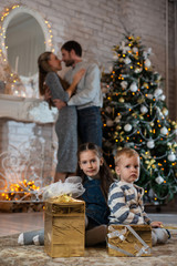Obraz na płótnie Canvas Sister hugs little brother sitting on the floor with gifts on background of their parents and Christmas tree