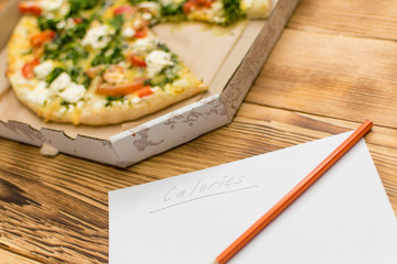 Count the calories. Sheet with the inscription Calories and lying near a pizza on a wooden background.
