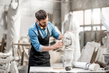 Handsome sculptor brushing stone head sculpture on the table in the atmospheric studio - obrazy, fototapety, plakaty