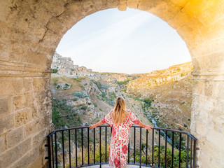 Young blonde girl looking out over the  landscape of the Sassi di Matera, prehistoric historic center, UNESCO World Heritage Site, European Capital of Culture 2019 (wide) - obrazy, fototapety, plakaty