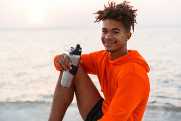 Cheerful young african guy outdoors on the beach drinking water.