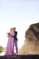 Beautiful young couple hugs and looks away and at each other enjoying a romantic date on the river. Man and woman standing on the rocks in the sun near the sea at sunset