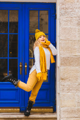 Blond curly romantic hipster woman near blue door of cozy cafe, resaurant or shop enjoying winter at snowy day. Female wearing white sweater, yelow knitted hat, sarf, gloves. City lifestyle concept.