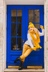 Blond curly romantic hipster woman near blue door of cozy cafe, resaurant or shop enjoying winter at snowy day. Female wearing white sweater, yelow knitted hat, sarf, gloves. City lifestyle concept.