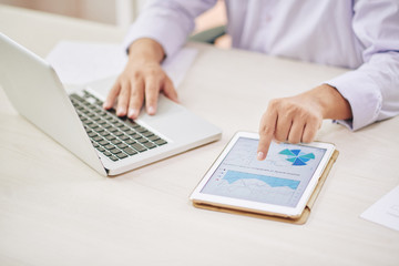 Fototapeta premium Faceless shot of male worker using laptop and tapping tablet with graphs