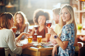 Woman looking at camera and holding glass of wine while sitting in restaurant. In background friends drinking and chatting.