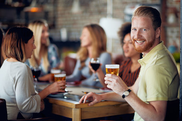 Man looking at camera and drinking beer while sitting in restaurant. In background friends chatting.