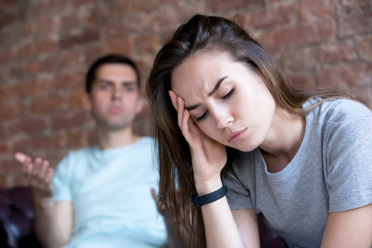 Offended man and woman are seated separately on sofa. Young couple in quarrel. Boyfriend and girlfriend fighting. Young stressed woman with headache, emotional arguing man. Family problems concept.