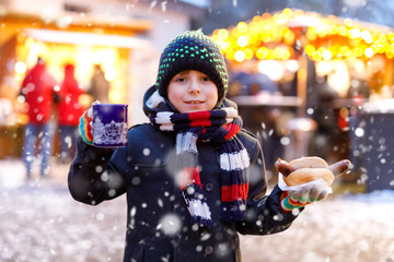 Little cute kid boy eating German sausage and drinking hot children punch on Christmas market....