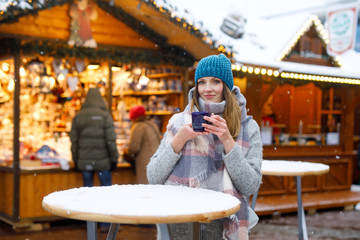 Beautiful young woman drinking hot punch, mulled wine on German Christmas market. Happy girl in winter clothes with lights on background on winter snow day in Munich, Germany.
