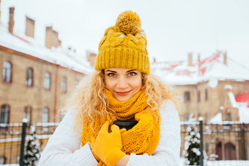 Outdoor portrait of blond curly woman in yellow hat, scarpf and gloves warming, drinking hot coffee or tea outdoor in winter city park square at snowy cold day.