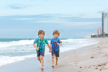 Two happy little kids boys running on the beach of ocean. Funny cute children, sibling and best friends making vacations and enjoying summer on stormy windy day. Healthy kids on beach of Miami, USA
