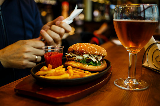 Burger with French fries and Beer, fork and knife in male hands on wooden table background