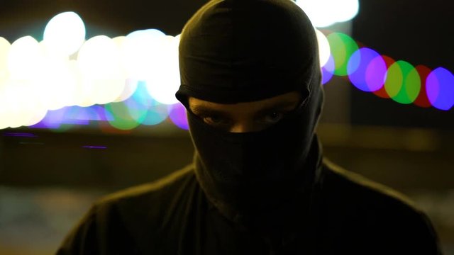 A man dressed in dark clothes and a balaclava goes with a bat in an abandoned park. There is an early winter outside and garlands hang around. His face is not visible in backlight. He is set up to fig