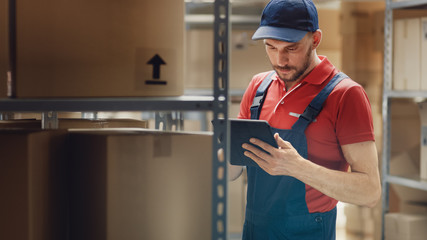 Handsome Warehouse Worker Uses Digital Tablet For Checking Stock, On the Shelves Standing Cardboard...