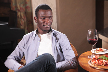 Young African American at the table in the restaurant with a glass of red wine and snacks. The concept of rest and communication. Photo in interior. Business lunch.