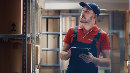Warehouse Worker Uses Digital Tablet For Checking Stock, On the Shelves Standing Cardboard Boxes.