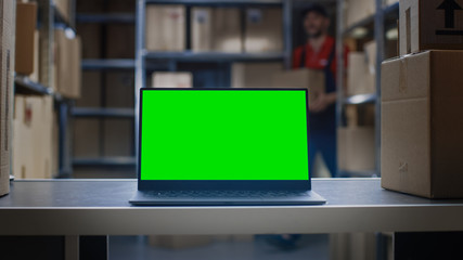 Laptop with Green Mock-up Screen Standing on the Desktop in the Warehouse where Man Works.