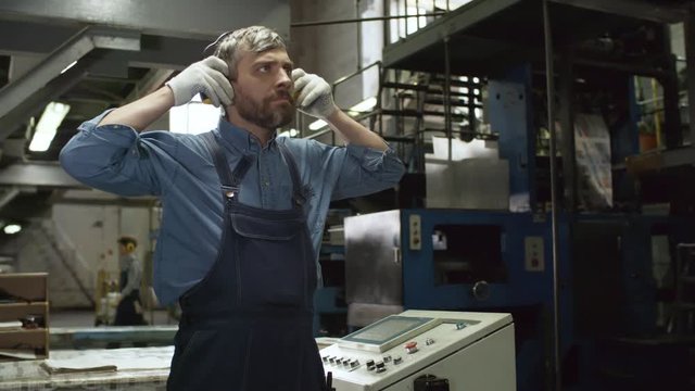Panning shot of middle aged Caucasian male technician putting on safety ear muffs, looking at industrial machine and pressing buttons on operating console while working in factory
