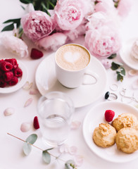 Delicious morning breakfast table essentials: fresh cappuccino coffee, marshmallow, cookies, chocolate, red raspberries, glass of water and tender pink blossoming peony bouquet on the white background