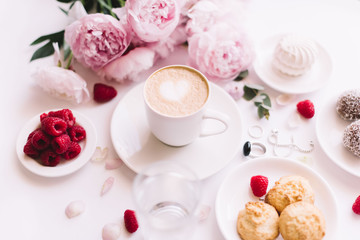Obraz na płótnie Canvas Delicious morning breakfast table essentials: fresh cappuccino coffee, marshmallow, cookies, chocolate, red raspberries, glass of water and tender pink blossoming peony bouquet on the white background