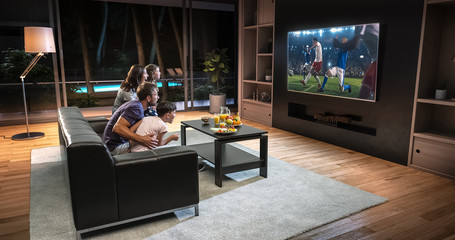 A family is watching a soccer moment on the TV and celebrating a goal, sitting on the couch in the living room. - obrazy, fototapety, plakaty