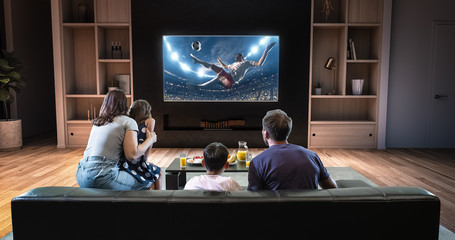 A family is watching a soccer moment on the TV and celebrating a goal, sitting on the couch in the living room. - obrazy, fototapety, plakaty