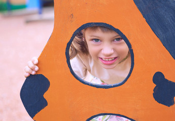 Face of small Caucasian girl looking through a hole in a play equipment outdoors. Orange background. Happy childhood concept