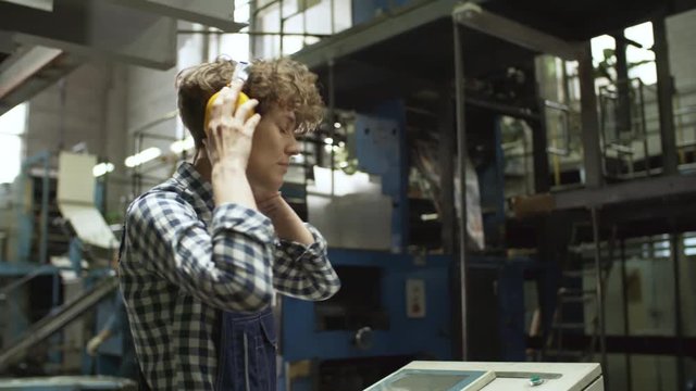 Young female technician putting on safety ear muffs and pressing buttons on operating console of industrial machine while working in plant