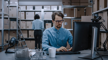 Fototapeta premium Warehouse Inventory Manager Works on Computer while Sitting at His Desk, In Background, Female Worker Check Shelf for a Delivery Package. Shelves are Full of Cardboard Box Packages Ready For Shipping.
