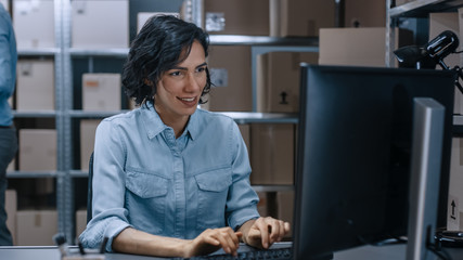 Warehouse Female Inventory Manager Works on a Computer while Sitting at Her Desk, In the Background...