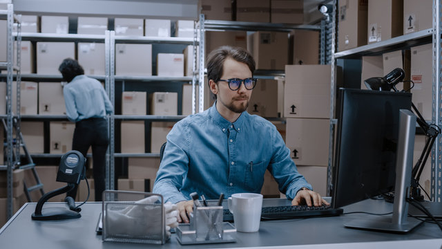 Warehouse Inventory Manager Works on Computer while Sitting at His Desk, In Background, Female Worker Check Shelf for a Delivery Package. Shelves are Full of Cardboard Box Packages Ready For Shipping.