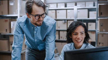 Male and Female Warehouse Inventory Managers Talking, Solving Problems, Using Personal Computer and Checking Stock. In the Background Rows of Shelves Full of Cardboard Box Packages.
