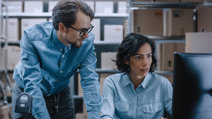 Male and Female Warehouse Inventory Managers Talking, Solving Problems, Using Personal Computer and Checking Stock. In the Background Rows of Shelves Full of Cardboard Box Packages.