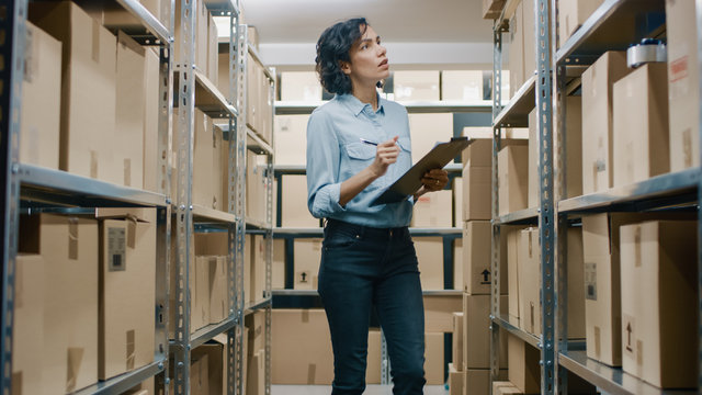 Female Inventory Manager Checks Stock, Writing in the Clipboard. Beautiful Woman Working in a Warehouse Storeroom with Rows of Shelves Full Of Cardboard Boxes, Parcels, Packages Ready for Shipment.