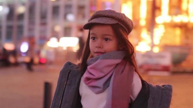 Child girl in a coat, cap and scarf is walking at the street against background of passing by people and evening city lights