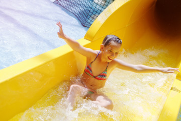 Young pretty girl in a striped colorful swimsuit laughs, rides on a yellow water slide in aqua park.
